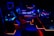 A high-tech music production studio illuminated by neon blue and red lights. Various equipment including keyboards, computer monitors, large speakers, and mixing consoles are present. An ergonomic office chair is positioned in front of the central monitoring station.