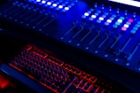 A dimly lit sound or lighting control board with multiple sliders and buttons, illuminated primarily by blue and red LED lights. Below it, a keyboard with glowing red backlighting provides a striking contrast.