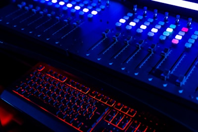A dimly lit sound or lighting control board with multiple sliders and buttons, illuminated primarily by blue and red LED lights. Below it, a keyboard with glowing red backlighting provides a striking contrast.