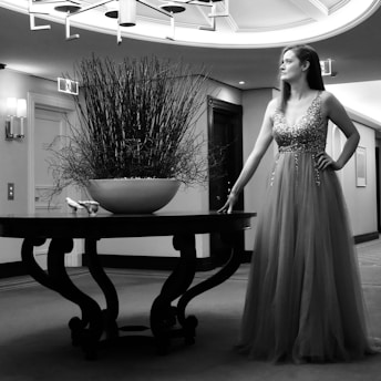 A woman in a sparkling evening gown poses next to a round wooden table. The table holds a large bowl filled with branches, creating a striking centerpiece. The setting appears to be an elegantly furnished corridor or hallway, with soft lighting and framed art on the walls. The atmosphere is sophisticated and serene.