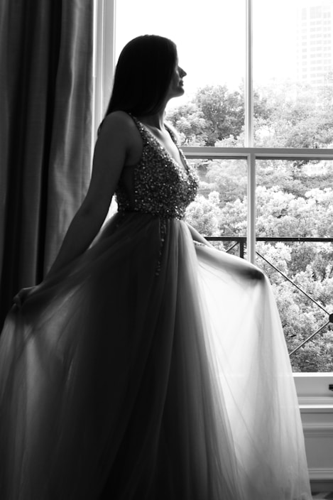 A graceful woman wearing a flowing deep blue evening gown standing by a softly lit window.