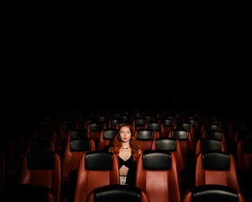 A lone person with red hair sits in the middle of a dimly lit, empty theater. Rows of maroon and black chairs surround them, creating a sense of solitude.