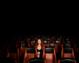 A lone person with red hair sits in the middle of a dimly lit, empty theater. Rows of maroon and black chairs surround them, creating a sense of solitude.
