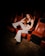 A woman with red hair is sitting in a theater chair, wearing a black top and black-and-white striped pants. The lighting casts a dramatic shadow around her, creating a focused spotlight effect. Her pose is relaxed, with one hand on her head and the other on the armrest.