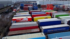 A large industrial storage yard filled with colorful shipping containers and trailers. The containers are stacked and aligned in rows, displaying various brand logos and colors. There are few vehicles parked nearby, and the area seems to be used for logistics or transportation purposes.