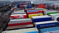 A large industrial storage yard filled with colorful shipping containers and trailers. The containers are stacked and aligned in rows, displaying various brand logos and colors. There are few vehicles parked nearby, and the area seems to be used for logistics or transportation purposes.