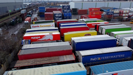 A large industrial storage yard filled with colorful shipping containers and trailers. The containers are stacked and aligned in rows, displaying various brand logos and colors. There are few vehicles parked nearby, and the area seems to be used for logistics or transportation purposes.