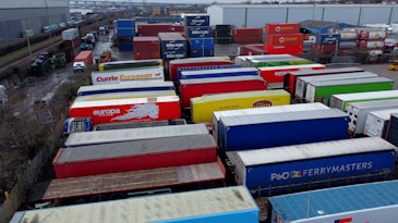A large industrial storage yard filled with colorful shipping containers and trailers. The containers are stacked and aligned in rows, displaying various brand logos and colors. There are few vehicles parked nearby, and the area seems to be used for logistics or transportation purposes.