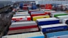 A large industrial storage yard filled with colorful shipping containers and trailers. The containers are stacked and aligned in rows, displaying various brand logos and colors. There are few vehicles parked nearby, and the area seems to be used for logistics or transportation purposes.