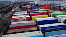 A large industrial storage yard filled with colorful shipping containers and trailers. The containers are stacked and aligned in rows, displaying various brand logos and colors. There are few vehicles parked nearby, and the area seems to be used for logistics or transportation purposes.