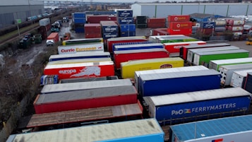A large industrial storage yard filled with colorful shipping containers and trailers. The containers are stacked and aligned in rows, displaying various brand logos and colors. There are few vehicles parked nearby, and the area seems to be used for logistics or transportation purposes.