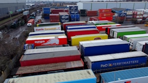 A large industrial storage yard filled with colorful shipping containers and trailers. The containers are stacked and aligned in rows, displaying various brand logos and colors. There are few vehicles parked nearby, and the area seems to be used for logistics or transportation purposes.