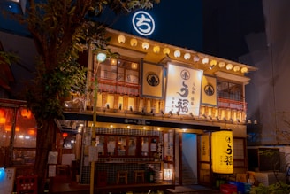 A traditional Japanese restaurant is brightly illuminated with warm lighting and lanterns, featuring kanji signage and a tiled exterior. The wooden structure has two stories, with a tree beside the building adding to the ambiance.