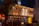 A traditional Japanese restaurant is brightly illuminated with warm lighting and lanterns, featuring kanji signage and a tiled exterior. The wooden structure has two stories, with a tree beside the building adding to the ambiance.