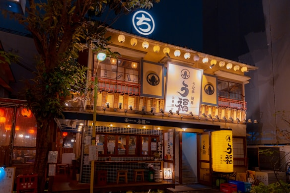 A traditional Japanese restaurant is brightly illuminated with warm lighting and lanterns, featuring kanji signage and a tiled exterior. The wooden structure has two stories, with a tree beside the building adding to the ambiance.