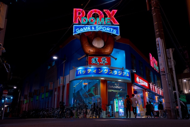 A vibrant, neon-lit facade of a gaming and sports establishment at night, featuring a large glove holding a baseball above the entrance. The building is adorned with colorful signs in both English and Japanese, with various advertisements and an illuminated sign that says 'ROX DOME'. Several people are walking towards the entrance, and bicycles are parked along the side.