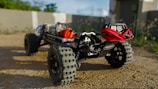 A close-up view of a rugged off-road vehicle with large, knobby tires on a dirt surface. The chassis reveals mechanical components, including a red coil or wire wrapped around the front bumper area. In the background, there are blurred structures and greenery under a clear sky.