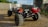 A close-up view of a rugged off-road vehicle with large, knobby tires on a dirt surface. The chassis reveals mechanical components, including a red coil or wire wrapped around the front bumper area. In the background, there are blurred structures and greenery under a clear sky.