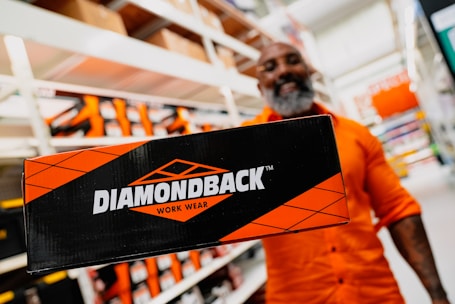 A person in an orange shirt is standing in an indoor setting, likely a store or warehouse, holding a black and orange box labeled 'Diamondback Work Wear'. Shelving with more boxes can be seen in the background.