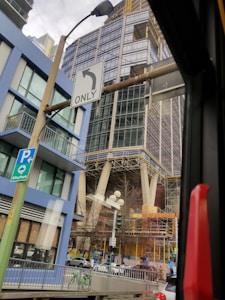 A tall modern building under construction is visible, surrounded by scaffolding and supported by large columns. In the foreground, there are road signs indicating directions, and a view of a city street with parked bicycles and cars. The scene has an urban atmosphere with a mix of construction materials and infrastructure.