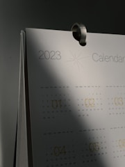 A close-up of a 2023 calendar with a metal hook at the top. The calendar pages show months with printed dates and a subtle starburst graphic near the year. Shadows cast a gradient effect over the surface, creating a sense of depth.