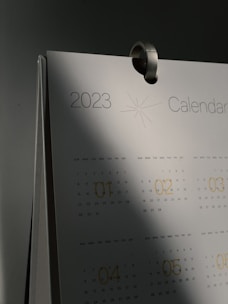A close-up of a 2023 calendar with a metal hook at the top. The calendar pages show months with printed dates and a subtle starburst graphic near the year. Shadows cast a gradient effect over the surface, creating a sense of depth.