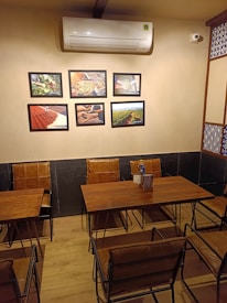 A small, cozy room with two wooden tables and cushioned chairs arranged neatly. On the cream-colored wall, there is an air conditioner above six framed photographs showcasing various scenes. The room has a warm, inviting look with a combination of traditional and modern decor elements.