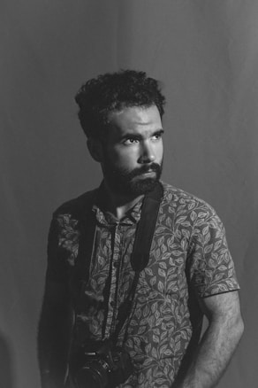 A man with a beard, wearing a patterned shirt, stands holding a camera around his neck. The image is in black and white, focusing on his contemplative expression, with soft lighting creating shadows.