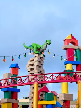 A large green dinosaur model is perched on top of a precariously stacked tower of Jenga blocks. Surrounding the tower are colorful building blocks in various shapes and sizes, some resembling Tinkertoy pieces. A string of oversized, colorful bulb lights drapes around the structures, and a red fence-like structure adds to the playful atmosphere. The sky is a clear blue, enhancing the vibrant colors of the scene.