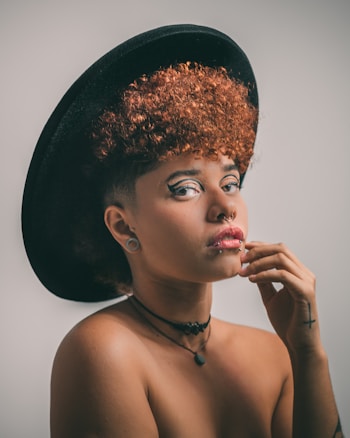 A person with vibrant curly hair is wearing a black wide-brimmed hat and multiple piercings, including a nose ring and lip piercing. They have artistic eyeliner and a contemplative expression, touching their chin lightly. A tattoo is visible on their hand, and they are adorned with necklaces.