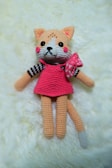 A crocheted plush toy resembling a cat with peach fur, a pink dress, and a bow on its side. The toy has large black eyes, a small black nose, and rosy cheeks. It is lying on a soft cream-colored surface.