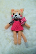 A crocheted plush toy resembling a cat with peach fur, a pink dress, and a bow on its side. The toy has large black eyes, a small black nose, and rosy cheeks. It is lying on a soft cream-colored surface.
