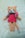 A crocheted plush toy resembling a cat with peach fur, a pink dress, and a bow on its side. The toy has large black eyes, a small black nose, and rosy cheeks. It is lying on a soft cream-colored surface.