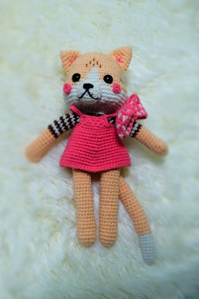 A crocheted plush toy resembling a cat with peach fur, a pink dress, and a bow on its side. The toy has large black eyes, a small black nose, and rosy cheeks. It is lying on a soft cream-colored surface.