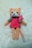 A crocheted plush toy resembling a cat with peach fur, a pink dress, and a bow on its side. The toy has large black eyes, a small black nose, and rosy cheeks. It is lying on a soft cream-colored surface.