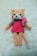A crocheted plush toy resembling a cat with peach fur, a pink dress, and a bow on its side. The toy has large black eyes, a small black nose, and rosy cheeks. It is lying on a soft cream-colored surface.