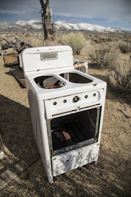 An abandoned, weathered stove sits in an arid, desert-like landscape. The appliance appears old and rusted, missing its burners and with a broken door. Surrounding it are dry bushes and a few scattered pieces of debris. In the background are snow-capped mountains under a clear blue sky.