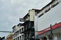 An urban street scene featuring a series of multi-story buildings with worn exteriors. The most prominent building displays a sign that reads 'The Guest.' Telephone wires and cables are visible in the foreground, adding to the urban atmosphere. The sky appears overcast, creating a muted lighting effect.