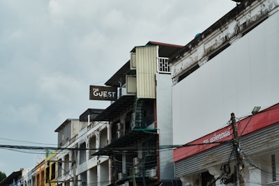 An urban street scene featuring a series of multi-story buildings with worn exteriors. The most prominent building displays a sign that reads 'The Guest.' Telephone wires and cables are visible in the foreground, adding to the urban atmosphere. The sky appears overcast, creating a muted lighting effect.