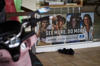 A storefront advertisement featuring a poster with images of four people wearing glasses, accompanied by the slogan 'SEE MORE. DO MORE.' in both English and another language. A pair of black shoes is placed on the tiled floor in front of the glass window. A pink scooter is partially visible on the left side of the image.