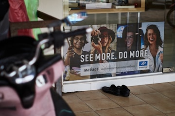 A storefront advertisement featuring a poster with images of four people wearing glasses, accompanied by the slogan 'SEE MORE. DO MORE.' in both English and another language. A pair of black shoes is placed on the tiled floor in front of the glass window. A pink scooter is partially visible on the left side of the image.