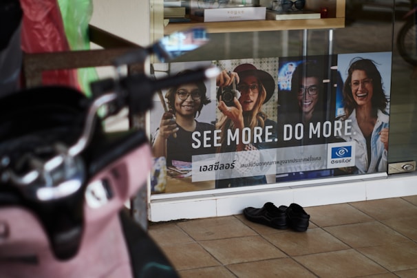 A storefront advertisement featuring a poster with images of four people wearing glasses, accompanied by the slogan 'SEE MORE. DO MORE.' in both English and another language. A pair of black shoes is placed on the tiled floor in front of the glass window. A pink scooter is partially visible on the left side of the image.