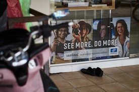 A storefront advertisement featuring a poster with images of four people wearing glasses, accompanied by the slogan 'SEE MORE. DO MORE.' in both English and another language. A pair of black shoes is placed on the tiled floor in front of the glass window. A pink scooter is partially visible on the left side of the image.