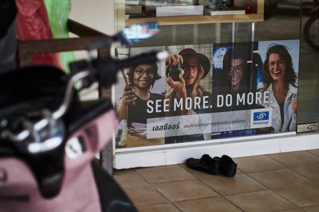 A storefront advertisement featuring a poster with images of four people wearing glasses, accompanied by the slogan 'SEE MORE. DO MORE.' in both English and another language. A pair of black shoes is placed on the tiled floor in front of the glass window. A pink scooter is partially visible on the left side of the image.