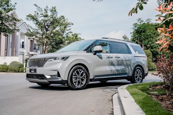 A sleek, modern minivan is parked on a neighborhood street surrounded by greenery and residential homes. The vehicle is metallic silver with stylish black alloy wheels and large windows. Trees and manicured bushes line the street, providing a serene suburban setting.