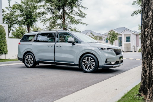 A modern minivan with a sleek, silver exterior is parked on a paved street. Surrounding the vehicle are well-maintained greenery and several contemporary suburban houses. The scene is well-lit, suggesting a daytime setting with a partly cloudy sky.