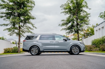 A sleek gray minivan is parked on a suburban street, flanked by two tall trees. The street is lined with greenery and well-maintained shrubs, against the backdrop of a cloudy sky. The minivan is centrally positioned, showcasing its side profile and modern design.