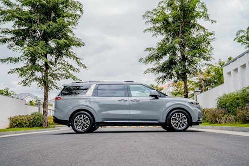 A sleek gray minivan is parked on a suburban street, flanked by two tall trees. The street is lined with greenery and well-maintained shrubs, against the backdrop of a cloudy sky. The minivan is centrally positioned, showcasing its side profile and modern design.