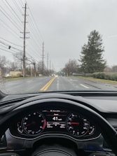 The image captures a view from the interior of a car looking out towards a street. The dashboard is visible, showing a speed of 41 mph. The weather appears overcast with a wet road, suggesting recent rain. There are power lines and trees lining the street.