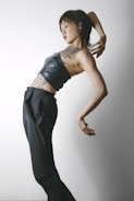 A person is posing artistically against a plain background. They are wearing a black leather top and high-waisted black trousers. Their torso is adorned with text, possibly resembling tattoos or body art. The posture is expressive and dynamic, suggesting movement or dance.